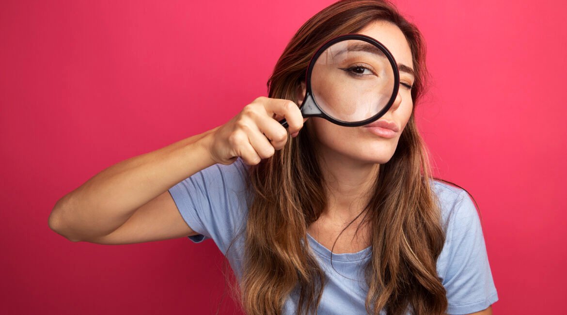 Jovem mulher bonita em camiseta azul olhando para a câmera através de lupa com interesse em pé sobre fundo rosa